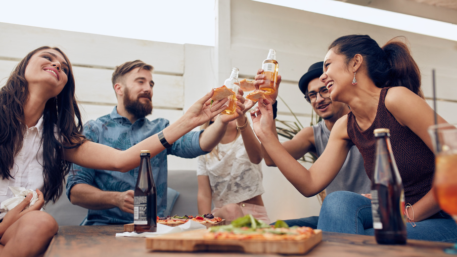Group of friends toasting beers in a party while sitting around table. Multiracial friends hanging out on rooftop enjoying party.