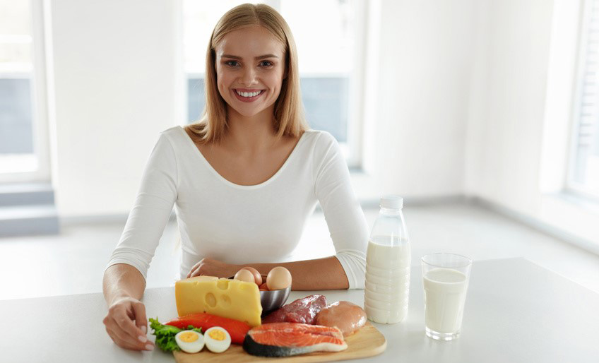 Healthy Nutrition. Beautiful Smiling Woman At Table With Different Food Products On It, Meat, Salmon, Chicken, Eggs, Cheese, Glass Of Milk On Kitchen Counter Top. Balanced Diet Foods. High Resolution