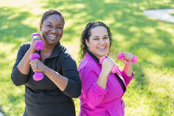 Two multi-ethnic women in their 30s exercising outdoors on a sunny day, lifting handweights. They are standing, smiling and looking at the camera.