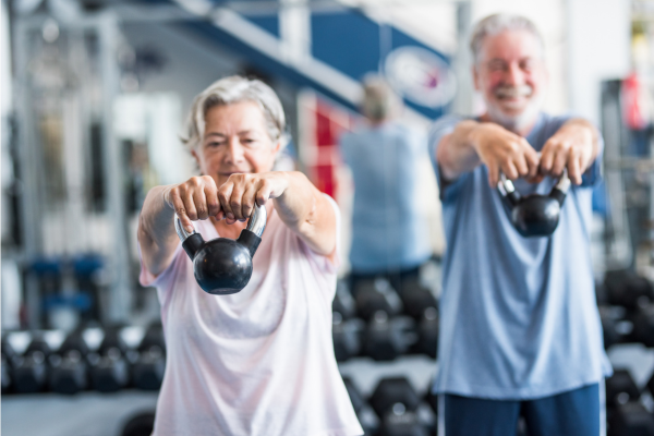 Older adult man and woman lifting kettlebells in a gym, demonstrating strength training and healthy aging"