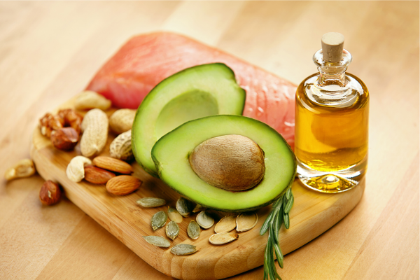 Assortment of healthy fats including avocado, mixed nuts, salmon, and a bottle of olive oil on a wooden cutting board.