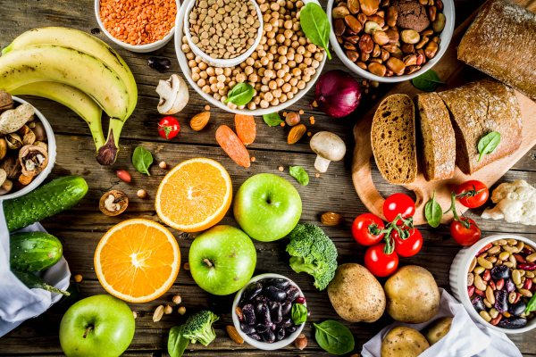 High-fiber foods including apples, oranges, bananas, legumes, lentils, whole grain bread, potatoes, and leafy greens displayed on a wooden table.