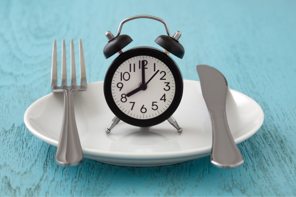 A small black alarm clock on a white plate with fork and knife, symbolizing meal timing or intermittent fasting.
