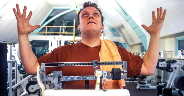 Man standing on a scale at the gym with arms raised in frustration, representing weight loss plateau